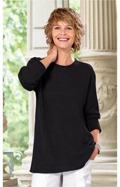 Schwarzer, langärmliger Pullover ohne Muster, mit rundem Halsausschnitt, locker geschnitten, getragen von lächelnder stehender Frau mit kurzen blonden Haaren, rechte Hand am Nacken, linke Hand in weißer Hose in Tasche