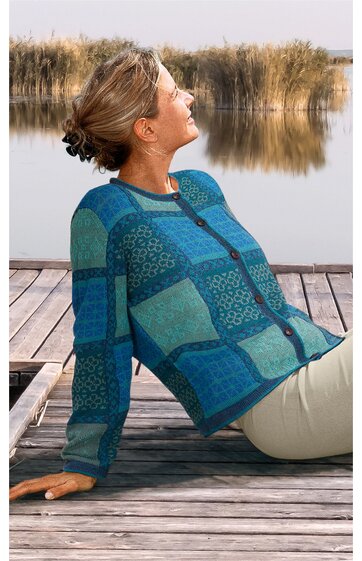 Frau sitzt entspannt auf einem Steg am Wasser, trägt einen blauen Pullover