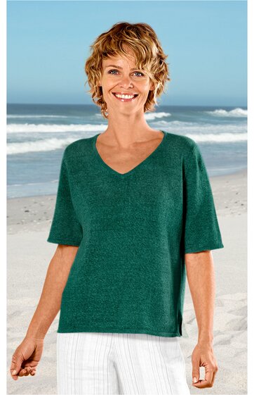 Damenpullover in dunklem Tannengrün, unifarben, V-Ausschnitt, kurze Ärmel, gerade geschnittener Saum mit kleinen seitlichen Schlitzen, getragen von lächelnder, stehender Frau am Strand