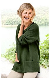 Damenpullover in dunkelgrün, einfarbig, langer Schnitt mit seitlichen Einschubtaschen, weiter Rundhalsausschnitt, getragen von lächelnder stehender Frau mit kurz blondem Haar