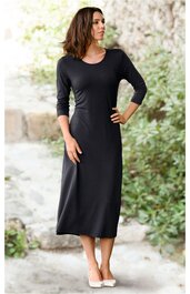 Schwarzes, knöchellanges Kleid mit 3/4-Ärmeln, rundem Ausschnitt, schmaler Passform, getragen von einer stehenden Frau mit linker Hand in der Hüfte und rechter Hand am Rock, schlichte, gerade Form