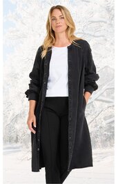 Schwarzer langer Mantel, Knopfleiste vorne, Stehkragen, lange Ärmel, getragen von stehender Frau, seitliche Taschen, offen getragen über weißem T-Shirt und schwarzer Hose