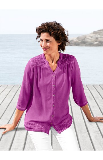 Bluse in magentaviolett, unifarben, mit Knopfleiste vorne, Stehkragen, 3/4-Ärmeln, Spitzenbesatz an Schultern und Saum, getragen von sitzender Frau am Holzsteg mit Blick aufs Wasser
