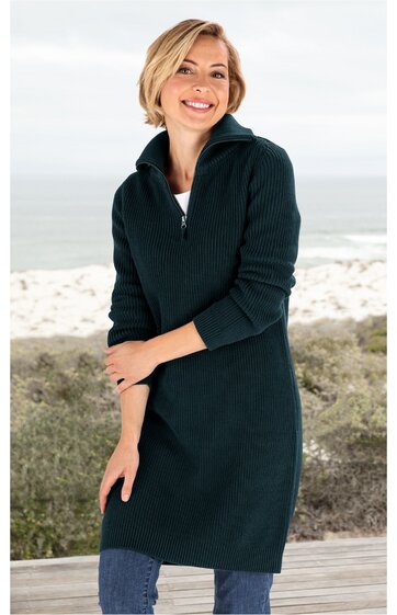 Dunkelgrüner Strickpullover-Kleid, langer Schnitt, Stehkragen mit Reißverschluss, geripptes Muster an Ärmeln und Kragen, lange Ärmel, getragen von lächelnder stehender Frau vor Strandkulisse