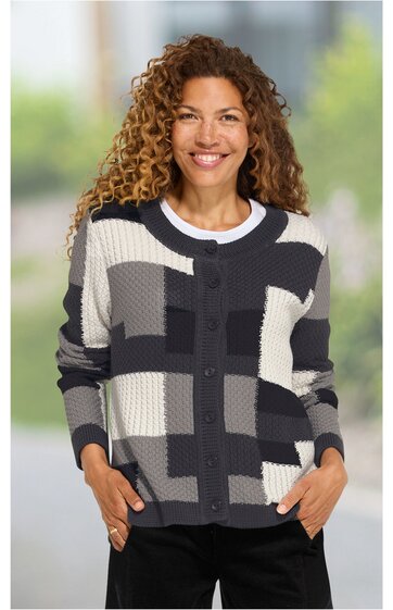 Strickjacke in Schwarz, Weiß und Grautönen mit großem geometrischem Karomuster, Rundhalsausschnitt, Knopfleiste, lange Ärmel, gerade geschnitten, getragen von lächelnder Frau in stehender Pose, Hände in den Taschen