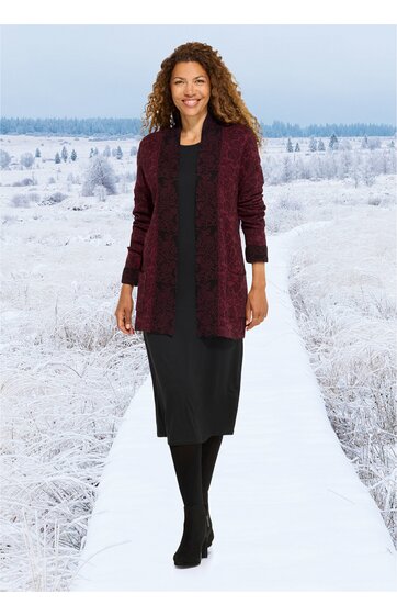 Langärmeliger bordeauxroter Cardigan mit offenem Schnitt, getragen von Frau mit lockigen Haaren, kombiniert mit schwarzem knielangem Kleid, schwarzer Strumpfhose und schwarzen Schuhen, stehende Pose auf weißem Weg vor schneebedeckter Landschaft