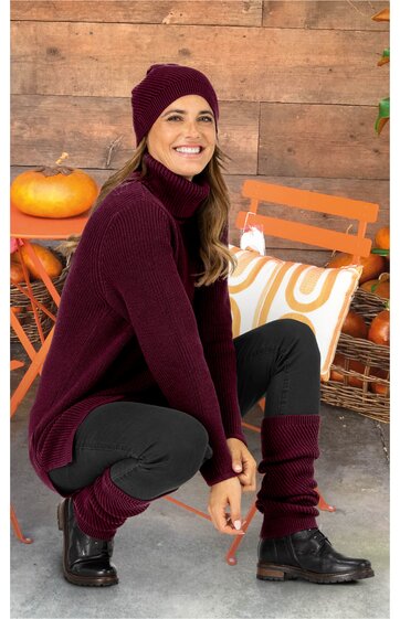 Dunkelroter Rollkragenpullover, schwarze Leggings, dunkelrote Mütze und Beinstulpen, schwarze Schnürstiefel, sitzende Frau, brauner Holzhintergrund mit Kürbissen und Korb