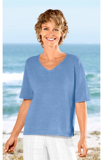 Hellblaues Kurzarmshirt mit V-Ausschnitt, gerade geschnitten, seitliche Schlitze am Saum, getragen von lächelnder Frau stehend vor Meereskulisse