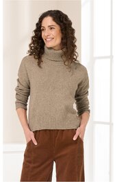 Beiger Rollkragenpullover, langärmlig, schlichter Schnitt, getragen von stehender Frau mit lockigen dunklen Haaren, entspannte Pose mit Händen in braunen Hosen gesteckt