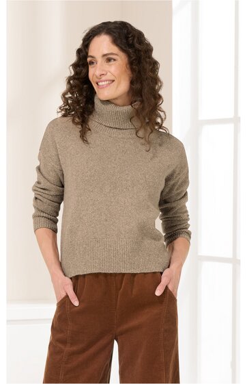 Beiger Rollkragenpullover, langärmlig, schlichter Schnitt, getragen von stehender Frau mit lockigen dunklen Haaren, entspannte Pose mit Händen in braunen Hosen gesteckt