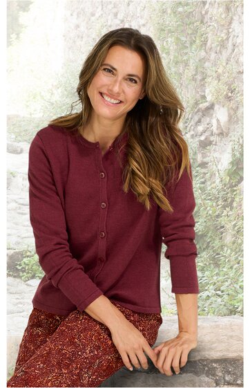 Bordeauxroter Damenstrickcardigan, langärmlig, rundhalsausschnitt, durchgeknöpft, feine Strickstruktur, getragen von sitzender Frau mit braunen, gemusterten Hosen, entspannte Pose