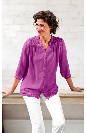 Langärmlige Bluse in violett mit Spitzenbesatz am Oberteil, durchgehender Knopfleiste, Stehkragen, locker sitzend, getragen von sitzender Frau mit weißen Hosen