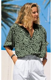 Damenbluse in dunklem Olivgrün mit hellgrünem Blumenmuster, V-Ausschnitt, kurze weite Ärmel, locker geschnitten, getragen von stehender Frau mit verschränkten Händen in weißen, hochgesetzen Hosen