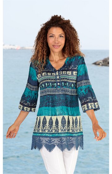 Tunika in türkis und blau mit indischem Muster, V-Ausschnitt, dreiviertellange Ärmel mit Volants, gerade geschnitten, Saum mit Spitzenbesatz, getragen von stehender Frau vor Meereshintergrund