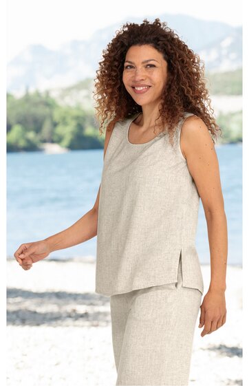 Ärmelloses, beiges Oberteil mit Rundhalsausschnitt und seitlichen Schlitzen, locker sitzend, getragen von stehender Frau vor Wasser- und Bergkulisse