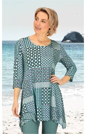 Längeres Damenshirt in Blau- und Grüntönen mit geometrischem Muster, Rundhalsausschnitt, dreiviertellange Ärmel, asymmetrischer Saum, getragen von stehender Frau am Strand