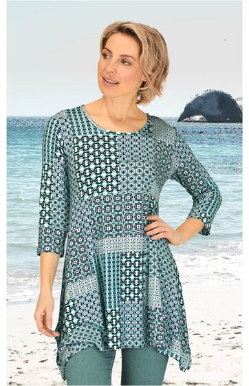 Längeres Damenshirt in Blau- und Grüntönen mit geometrischem Muster, Rundhalsausschnitt, dreiviertellange Ärmel, asymmetrischer Saum, getragen von stehender Frau am Strand