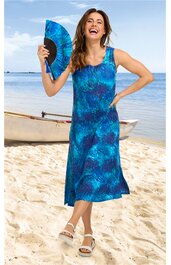 Ärmelloses, knielanges Sommerkleid in Blau mit abstraktem Muster, runder Ausschnitt, figurbetont, getragen von stehender Frau mit Handfächer, Sandstrand und Kanu im Hintergrund