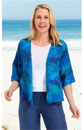Offene Strickjacke in Blau mit abstraktem Muster, halblange Ärmel, ohne Verschluss, getragen von lächelnder Frau stehend am Strand, kombiniert mit weißem T-Shirt und blauer Hose