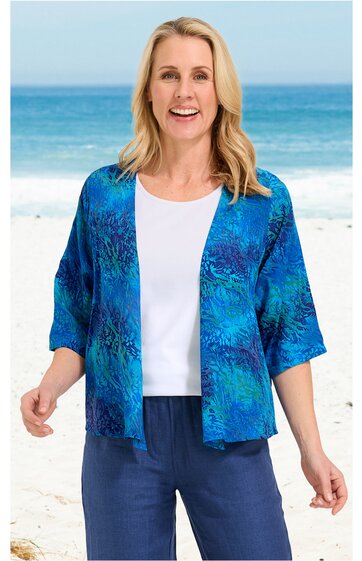 Offene Strickjacke in Blau mit abstraktem Muster, halblange Ärmel, ohne Verschluss, getragen von lächelnder Frau stehend am Strand, kombiniert mit weißem T-Shirt und blauer Hose