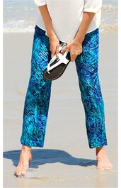 Badehose, türkis mit blauem und schwarzem abstraktem Muster, gerade geschnitten, getragen von stehender Person am Strand, mit nackten Füßen, Person hält weiße und schwarze Sandale in den Händen