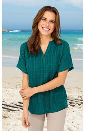 Locker geschnittene Bluse in dunklem Tannengrün mit feinem Karomuster, kurzer überschnittener Ärmel, V-Ausschnitt mit kleiner Knopfleiste, getragen von lächelnder Frau am Strand