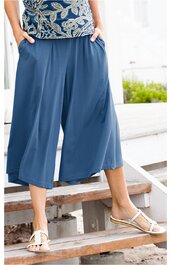 Weite kniehohe Hose in Stahlblau, glatter Stoff, elastischer Bund, gerade geschnitten, getragen von stehender Person mit Händen in den Taschen, kombiniert mit weißen Sandalen