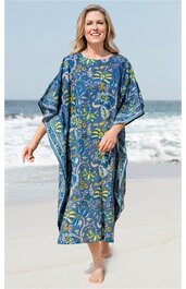 Blaues Kaftankleid mit floralen gelben und grünen Mustern, rundem Ausschnitt, weitem Schnitt, kurze Ärmel, getragen von lächelnder stehender Frau am Strand, knöchellang