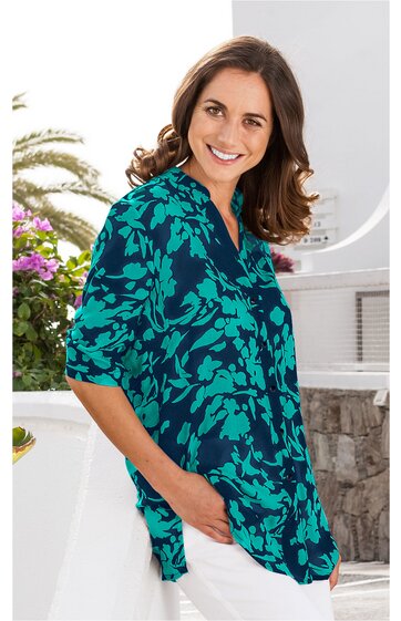 Türkisgrüne Bluse mit dunkelblauem Blumenmuster, V-Ausschnitt, Stehkragen, Knopfleiste, 3/4-Ärmel, locker sitzend, getragen von lächelnder Frau mit Händen in den Taschen, seitlich stehend