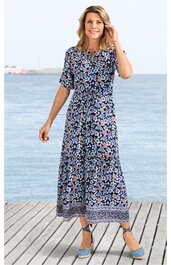 Maxikleid in Marineblau mit floralen Mustern in Hellblau, Rosa und Weiß, Rundhalsausschnitt, halblange Ärmel, gerade geschnittener Rock mit Bordürendruck, getragen von stehender lächelnder Frau am Wasser, knöchellang