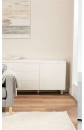 Weißes Sideboard mit vier Schubladen und einem Türfach, rechteckig, metallische Beine, steht auf Holzfußboden neben grauem Sofa mit Kissen und Decke, heller Raum mit Teppich und Wandbild