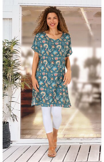 Blaues Kleid mit floralem Muster in Weiß und Gelbtönen, V-Ausschnitt, kurze Ärmel, knielang, gerade Form, getragen von stehender Frau auf Terrasse, kombiniert mit weißen Leggings