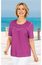 Damen-Shirt in fuchsia, halbtransparente Spitzeneinsätze am Rundhalsausschnitt und Ärmelenden, kurze Ärmel, lockere Passform, getragen von stehender Frau vor Meereskulisse