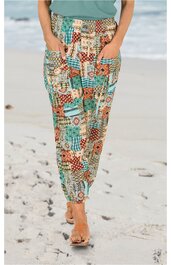 Locker sitzende lange Hose in Beige mit blau-rotem Patchwork-Muster, elastischer Bund, seitliche Eingrifftaschen, getragen von stehender Person am Strand