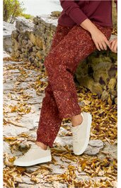 Rostrote Hose mit weißem abstraktem Muster, gerade geschnitten, getragen von sitzender Person, dazu weiße Schnürschuhe, herbstlicher Hintergrund mit Laub und Steinmauer