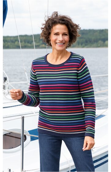 Langärmliges Shirt in dunklem Marineblau mit horizontalen Streifen in Rot, Hellblau, Weiß und Dunkelgrün, Rundhalsausschnitt, getragen von lächelnder stehender Frau vor Segelboot im Freien