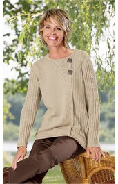 Beiger Pullover mit asymmetrischem Rippenstrickmuster auf einer Seite, Rundhalsausschnitt, zwei große dunkelgraue Knöpfe am Schulterbereich, lange Ärmel, getragen von sitzender Frau im Freien