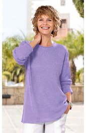 Hellvioletter Pullover mit Rundhalsausschnitt, lange Ärmel, lockere Passform, getragen von stehender lächelnder Frau, rechte Hand im Haar, linke Hand in Hosentasche, weißer Hintergrund mit Pflanzen