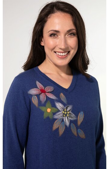 Blauer Damenpullover mit V-Ausschnitt und floralem Muster in Rosa, Gelb und Grau auf der linken Schulter, lange Ärmel, getragen von lächelnder stehender Frau