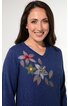 Blauer Damenpullover mit V-Ausschnitt und floralem Muster in Rosa, Gelb und Grau auf der linken Schulter, lange Ärmel, getragen von lächelnder stehender Frau