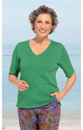 Grüner Pullover mit halblangen Ärmeln, V-Ausschnitt, gerade geschnitten, getragen von lächelnder Frau mit kurzen lockigen Haaren, stehend vor Meereshintergrund, keine weiteren sichtbaren Details