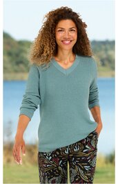Pullover in hellem Aquamarinblau, V-Ausschnitt, lange Ärmel, leicht lockere Passform, getragen von stehender Frau mit lockigen Haaren, kombiniert mit dunkler, gemusterter Hose mit Blumen- und Blätterdesign