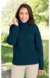 Dunkelgrüner Rollkragenpullover mit feiner Rippenstruktur, lange Ärmel, gerade geschnitten, getragen von Frau mit braunen Haaren, stehend, rechte Hand am Kragen, beige Hose