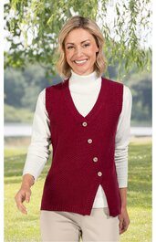 Damenstrickweste bordeauxrot, asymmetrische Knopfleiste mit vier Knöpfen, V-Ausschnitt, längere Passform, getragen von lächelnder stehender Frau mit weißen Langarmshirt und heller Hose, Outdoor-Hintergrund