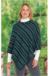 Poncho in dunkelgrün mit diagonalen Streifen in Blau, Grün und Beige, geometrisches Muster, asymmetrischer Saum, getragen von stehender Frau mit weißem Rollkragenpullover und grauer Hose