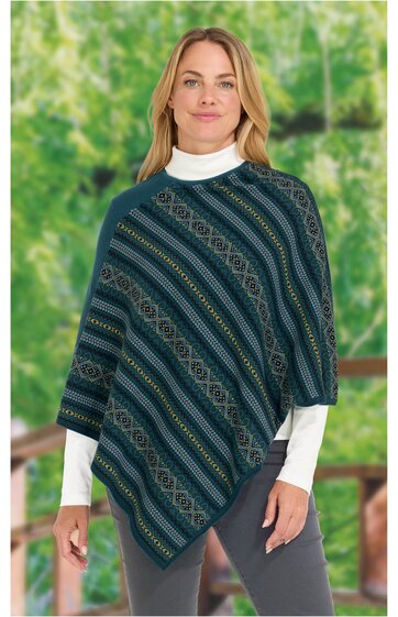Poncho in dunkelgrün mit diagonalen Streifen in Blau, Grün und Beige, geometrisches Muster, asymmetrischer Saum, getragen von stehender Frau mit weißem Rollkragenpullover und grauer Hose
