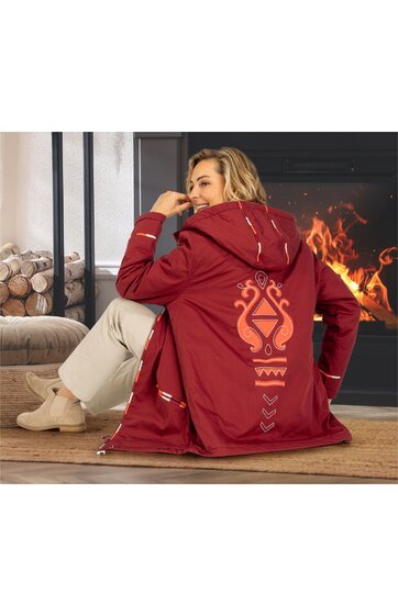 Rotes Damenjacke mit aufgesetzter Kapuze, großem orangefarbenem Muster auf dem Rücken, Reißverschluss, geschnittener Saum, getragen von sitzender Frau mit hellen Hosen und hellen Schuhen in Innenraum vor Kamin