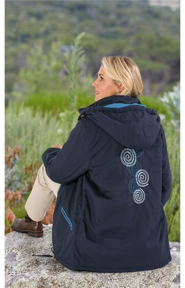 Damenjacke in Dunkelblau mit Kapuze, blaue Spiralstickereien auf dem Rücken, seitliche Eingrifftaschen mit hellblauen Reißverschlüssen, getragen von sitzender Person von hinten, sportiv und wetterfest