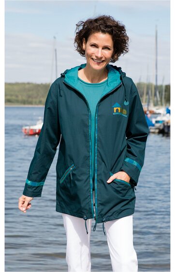 Dunkelgrüne Regenjacke mit Kapuze und Reißverschluss, türkisfarbene Ärmelbündchen und Innenseite der Kapuze, zwei Eingrifftaschen, taillierte Form, getragen von lächelnder Frau vor Wasser und Booten