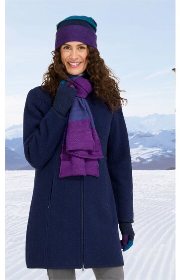 Marineblauer Mantel, langer Schnitt, durchgehender Reißverschluss, getragen von stehender Frau, dazu violett-blauer Schal, violette Mütze, marineblaue Handschuhe, winterliche Schneelandschaft im Hintergrund